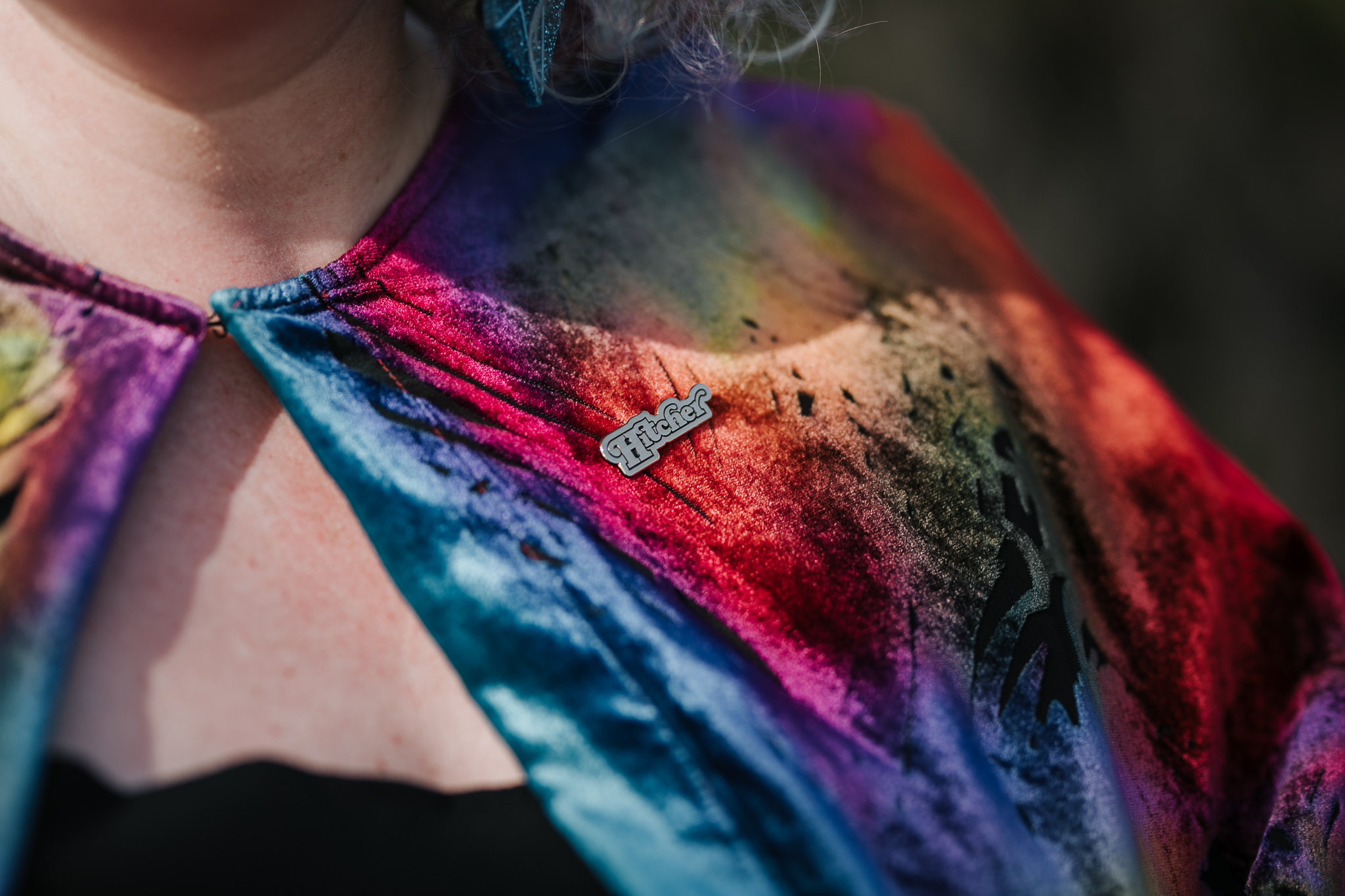 close up of a shoulder wearing a rainbow velvet cape and a golden pin that says "hitcher"