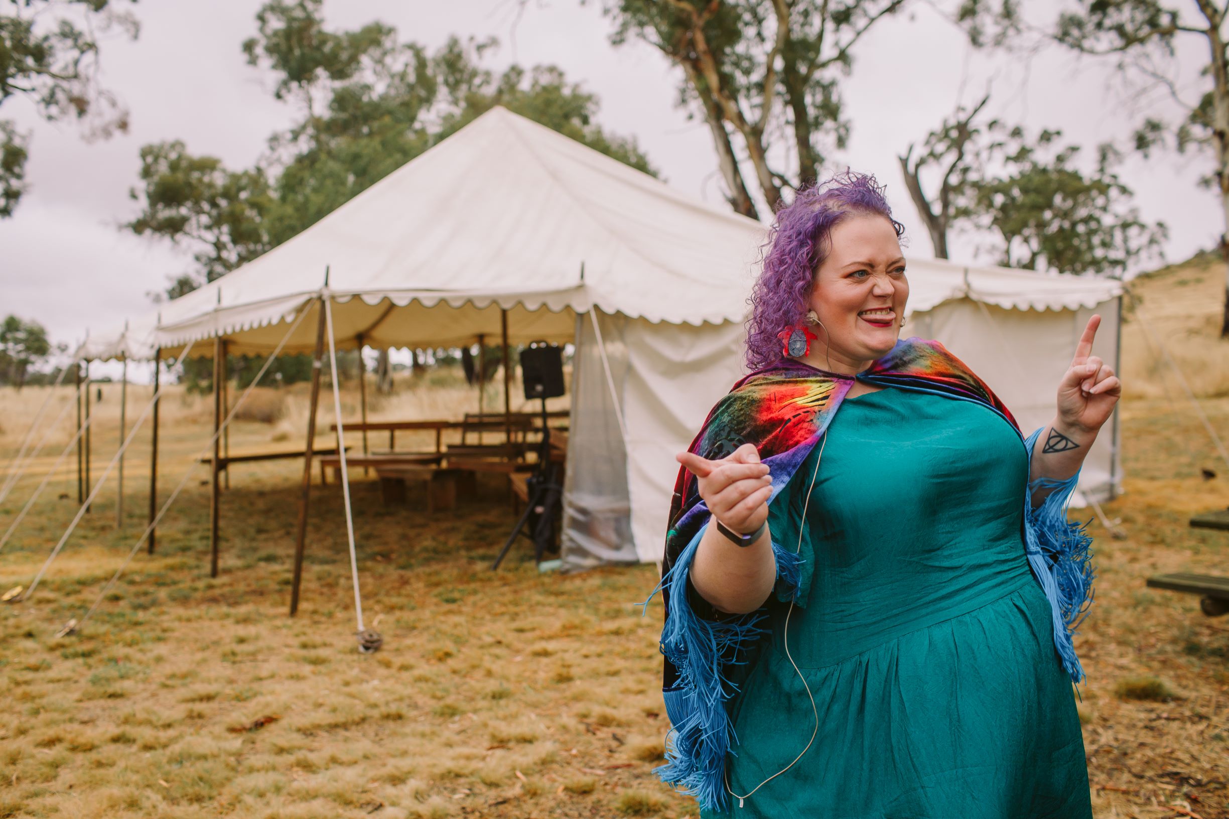 a person with curly purple hair in a colourful dress is having a little boogie in front of an empty marquee