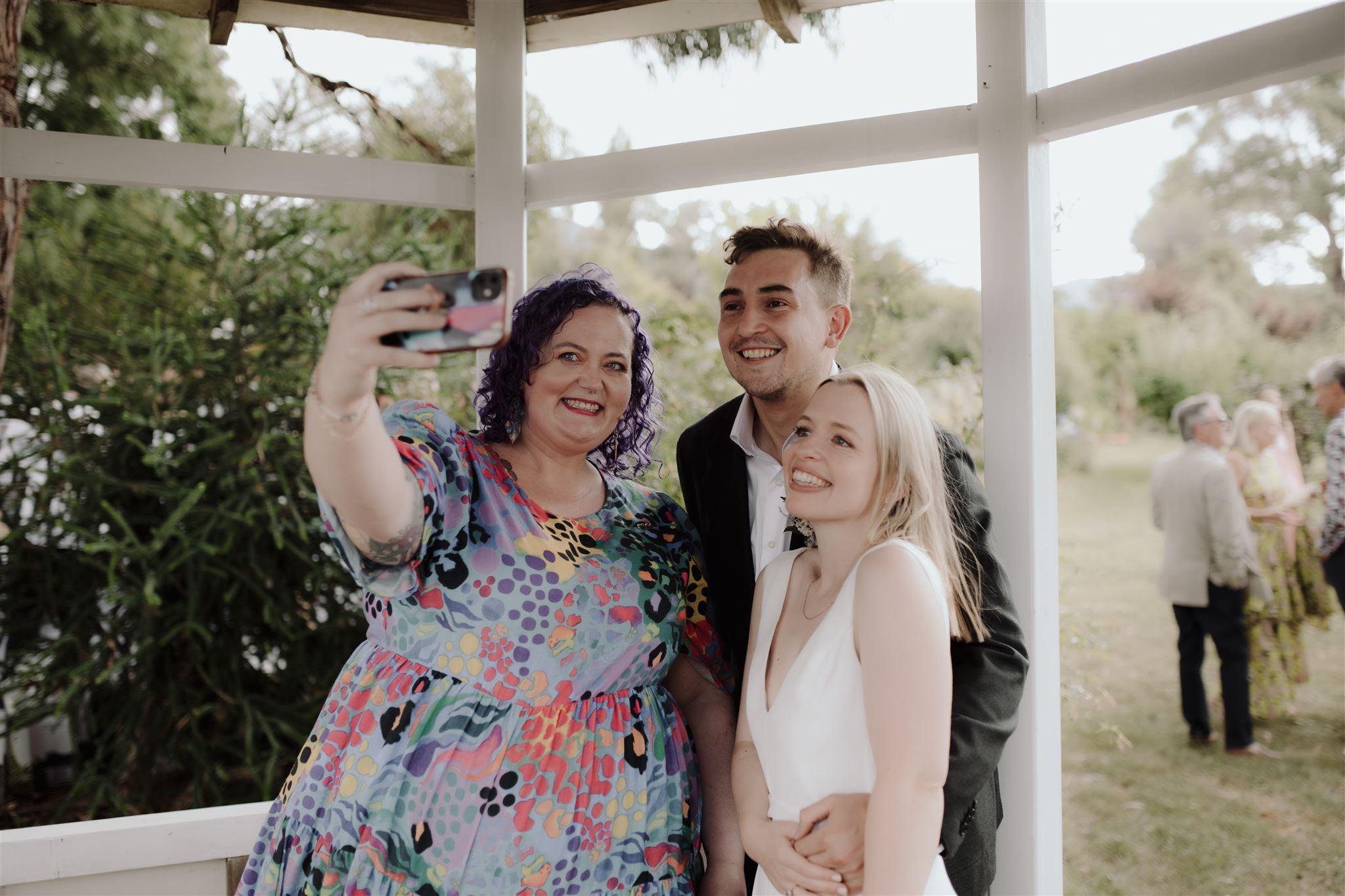 Three people are taking a selfie—two have just gotten married and the third person is holding the phone out in front of them all. 